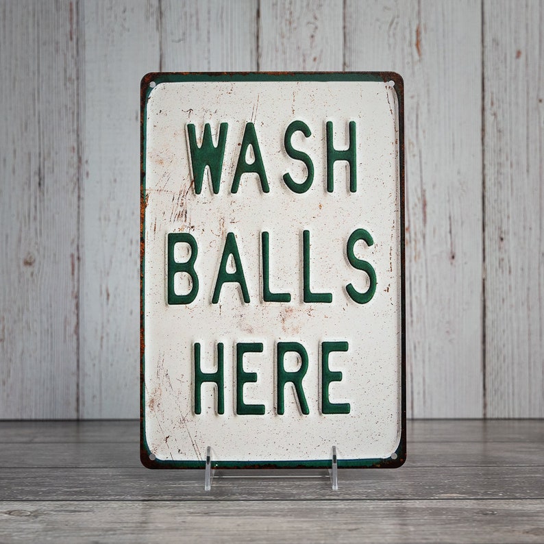 Peut inclure: Une pancarte en m&eacute;tal blanc avec des lettres vertes qui indiquent "WASH BALLS HERE". La pancarte a un aspect vieilli et est l&eacute;g&egrave;rement rouill&eacute;e.