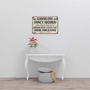 May include: A vintage-style metal sign with a white background and black text that reads "ALL GAMBLERS AND FANCY WOMEN MUST SIGN UP WITH CAPTAIN BEFORE BOAT LEAVES FOR NEW ORLEANS".