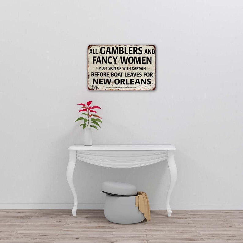 May include: A vintage-style metal sign with a white background and black text that reads "ALL GAMBLERS AND FANCY WOMEN MUST SIGN UP WITH CAPTAIN BEFORE BOAT LEAVES FOR NEW ORLEANS".