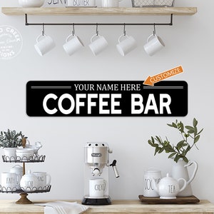 May include: Black and white sign with the text "YOUR NAME HERE COFFEE BAR" in a retro style. The sign is hanging on a white wall above a coffee bar setup with a coffee maker, mugs, and a sugar dispenser.