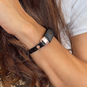 Può includere: Un braccialetto in pelle nera con chiusura in oro rosa. Il braccialetto è indossato al polso.