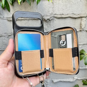 May include: Black leather phone case with a wallet design. The case has a zipper closure and includes card slots and a pocket. A black strap is attached for carrying. The interior features a tan-colored lining and a retro cassette tape design.