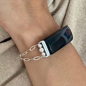 Può includere: Un tracker fitness nero con una catena argentata e perline bianche. Il tracker è indossato al polso.