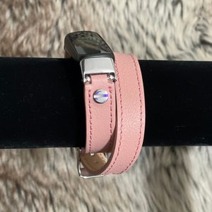Puede incluir: Una banda de doble vuelta de cuero rosa para un rastreador de fitness. La banda tiene una hebilla plateada y un acento plateado en la correa superior.