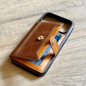 May include: Brown leather phone case with a card holder. The case has a magnetic closure and a contrasting stitch design. The card holder is visible, with a blue card partially showing. The case is on a wooden surface.