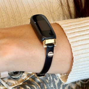 Op de afbeelding: Zwarte smartwatch met een rechthoekig scherm en goudkleurige accenten. De horlogeband is van zwart leer met een zilverkleurige studsluiting. Het horloge wordt om de pols gedragen.