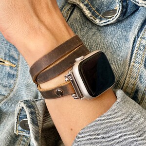 May include: A brown leather watch band wraps around a silver Apple Watch. The band is made of three separate straps that are stitched together.