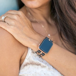 May include: A black leather watch strap with rose gold accents and studs. The strap is wrapped around a wrist and features a black smartwatch face.