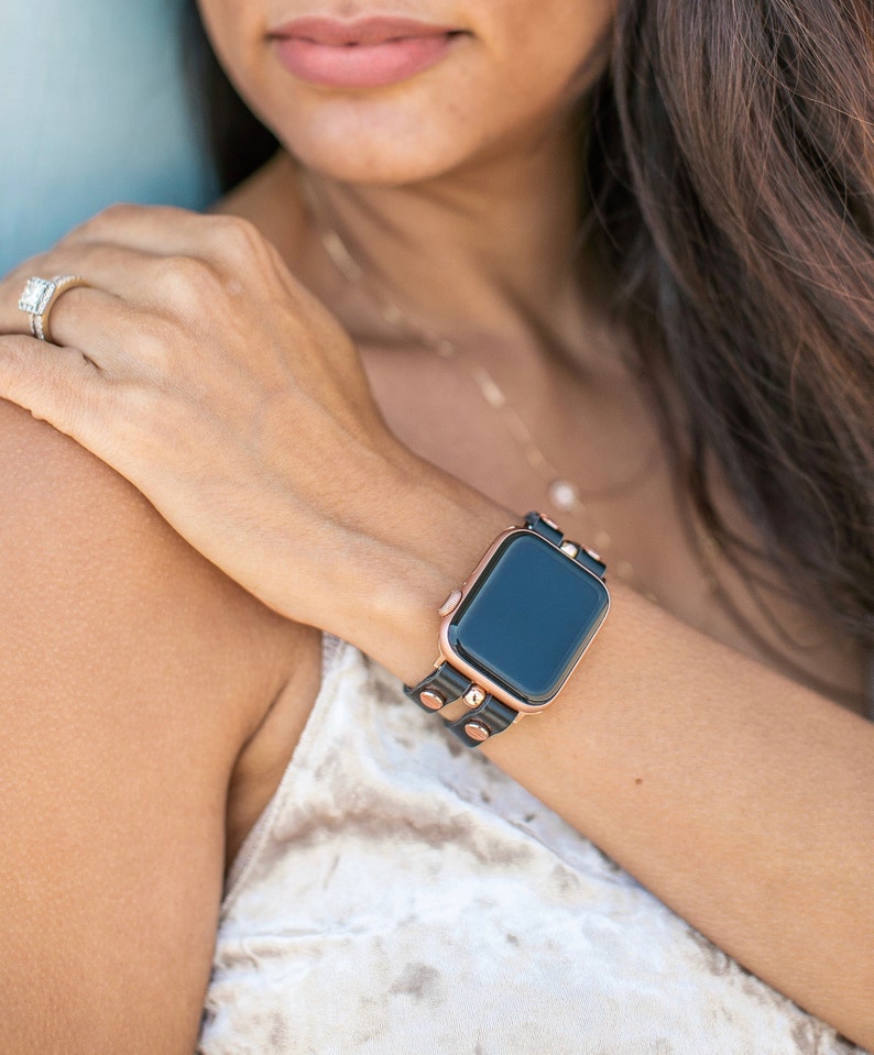 May include: A black leather watch strap with rose gold accents and studs. The strap is wrapped around a wrist and features a black smartwatch face.