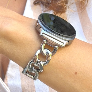 May include: A smartwatch with a dark, circular screen and a polished silver-tone metal chain link band. The watch features a metallic casing that connects to the interlocking, rounded links of the bracelet, creating a cohesive, reflective appearance.