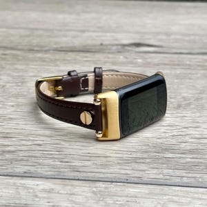 Op de afbeelding: Een bruin lederen horlogebandje met een goudkleurig frame en gesp. De wijzerplaat van het horloge is zwart. De band is gemaakt van donkerbruin leer met een lichtbruine binnenkant.