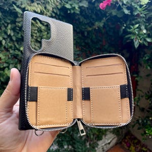 May include: A black leather phone case with a zippered wallet attached. The wallet has multiple card slots and a zippered compartment. The case is open and the interior is brown leather.