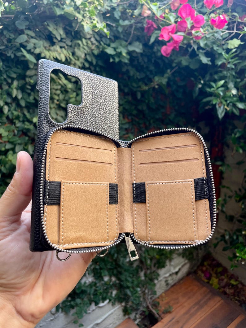 May include: A black leather phone case with a zippered wallet attached. The wallet has multiple card slots and a zippered compartment. The case is open and the interior is brown leather.