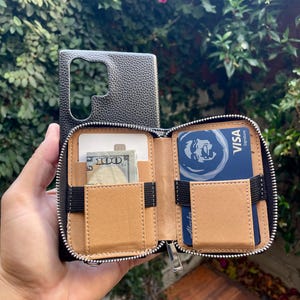 May include: A black leather phone case with a zippered wallet attachment. The wallet has two card slots, a cash pocket, and a zippered compartment. The case is open and shows the interior lining, which is a light brown color.