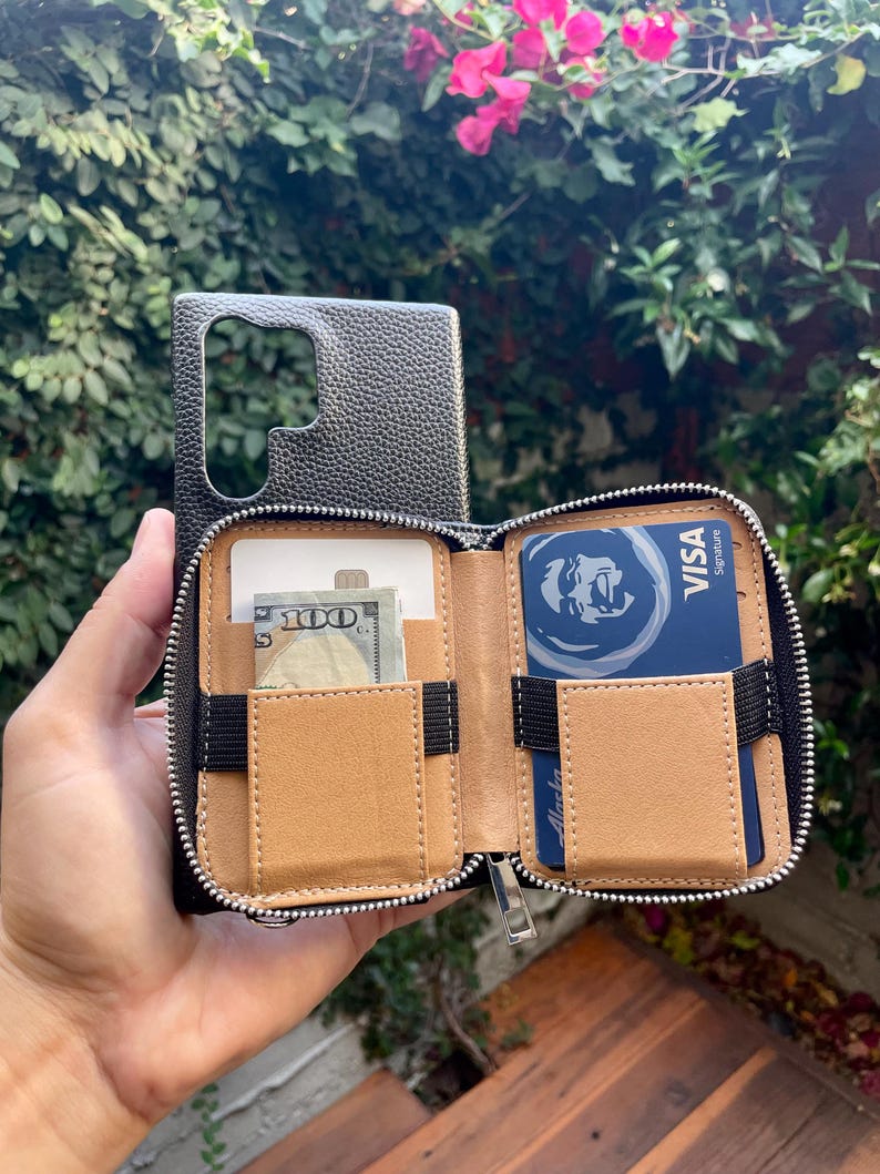 May include: A black leather phone case with a zippered wallet attachment. The wallet has two card slots, a cash pocket, and a zippered compartment. The case is open and shows the interior lining, which is a light brown color.