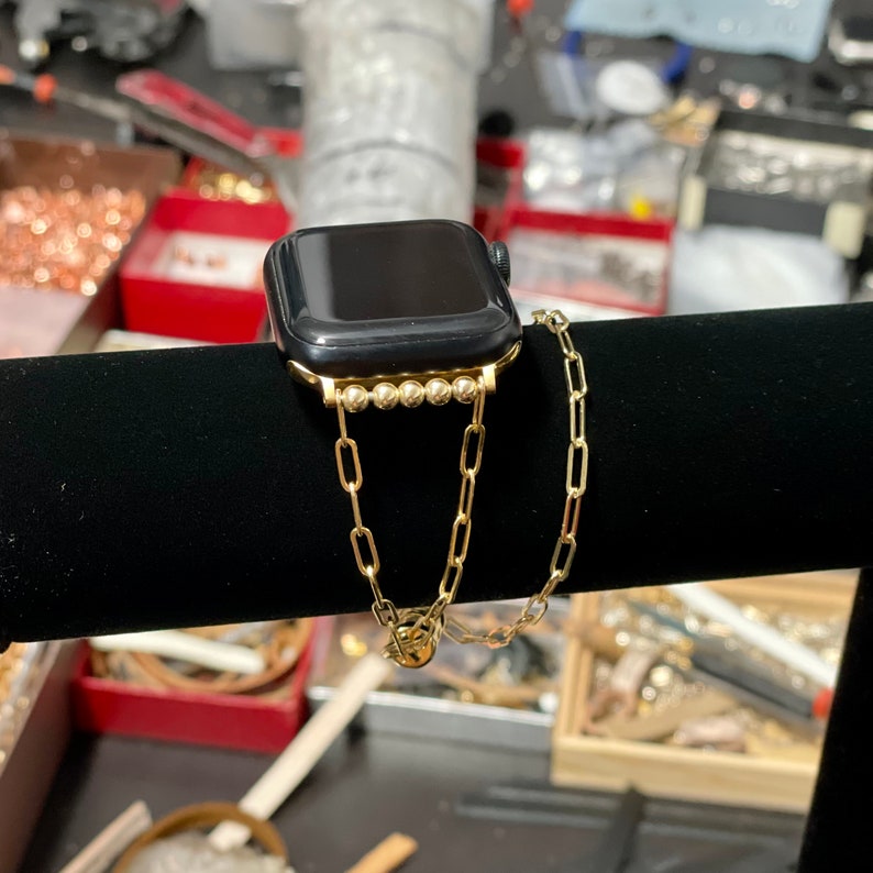 Puede incluir: Una pulsera de reloj de cadena dorada con una fila de peque&ntilde;as cuentas doradas. La pulsera est&aacute; unida a un Apple Watch negro.