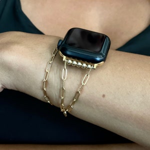 Puede incluir: Una pulsera de reloj de cadena dorada con un Apple Watch negro. La pulsera est&aacute; hecha de m&uacute;ltiples cadenas enlazadas.