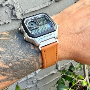 May include: A Casio Illuminator digital watch with a silver-tone rectangular case, a black bezel, and a tan leather strap. The watch face features a digital display, a circular analog-style sub-dial, and the text Casio, Illuminator, and WR100M.