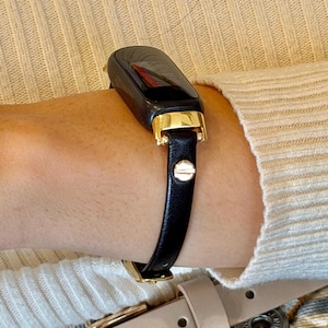 May include: Black and gold smartwatch with a black leather band. The watch has a rectangular face with a gold bezel and a black strap with a gold buckle. The watch is worn on the wrist.