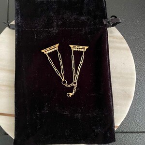 May include: Two gold-toned Apple Watch band connectors with a chain link design. The connectors are on a black velvet pouch.