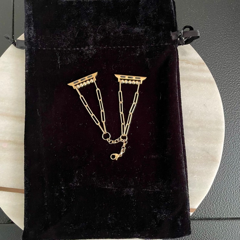 May include: Two gold-toned Apple Watch band connectors with a chain link design. The connectors are on a black velvet pouch.