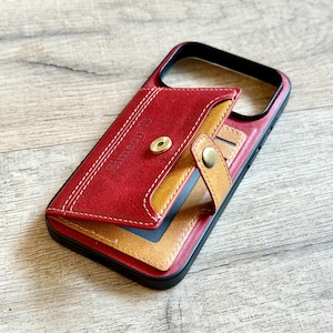Peut inclure: Étui de téléphone en cuir rouge et beige avec fonction portefeuille. L'étui a un cadre noir et une fermeture magnétique. La section portefeuille est beige et rouge avec le nom "Simeon D" cousu sur le devant.