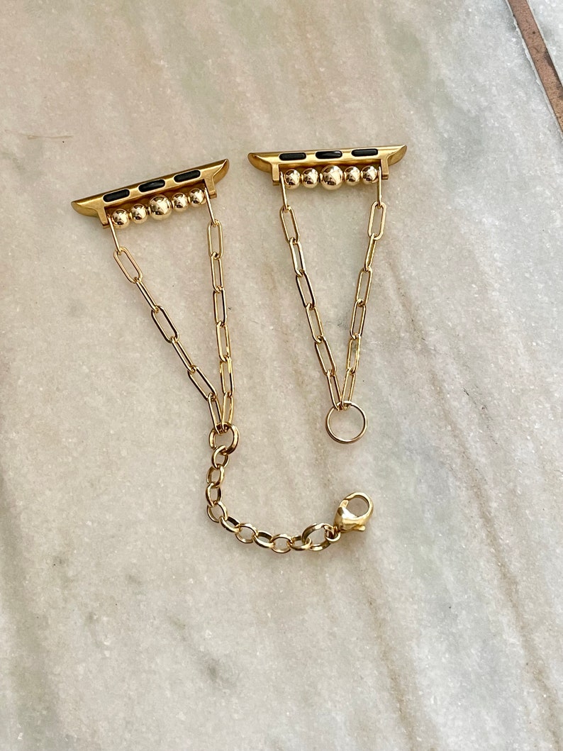 May include: Gold chain watch band with black accents and a clasp closure. The band is made of linked chains and has a decorative design.