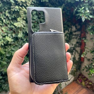 May include: Black leather phone case with a zippered wallet attached. The case has a textured finish and a cutout for the camera lens.