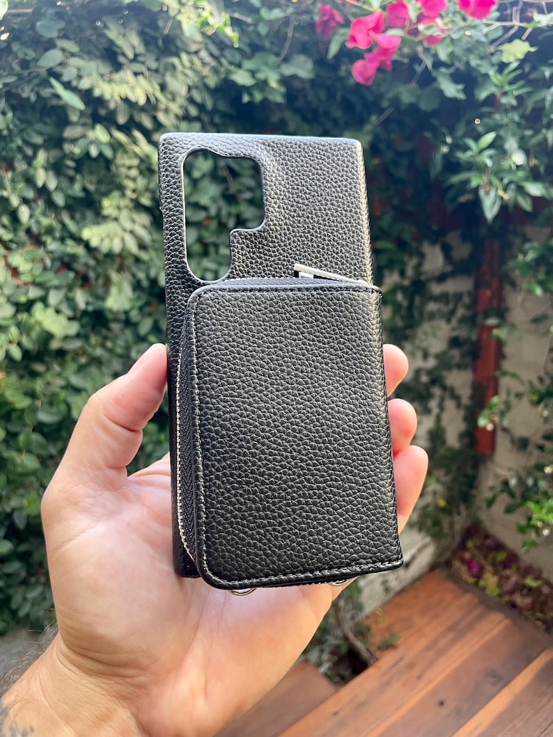 May include: Black leather phone case with a zippered wallet attached. The case has a textured finish and a cutout for the camera lens.