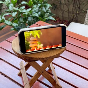 May include: A white phone case with a screen displaying a spa-like image of stacked stones, white flowers, and lit candles. The phone is sitting on a small wooden folding stool.