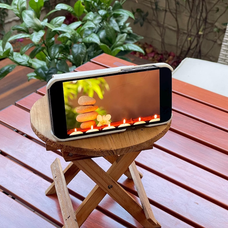 May include: A white phone case with a screen displaying a spa-like image of stacked stones, white flowers, and lit candles. The phone is sitting on a small wooden folding stool.