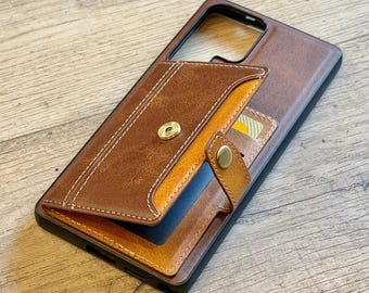 Op de afbeelding: Een bruine leren telefoonhoes met een kaarthouder. De hoes heeft een magnetische sluiting en een contrasterend oranje interieur. De stiksels zijn wit. Een creditcard is zichtbaar in de kaarthouder.