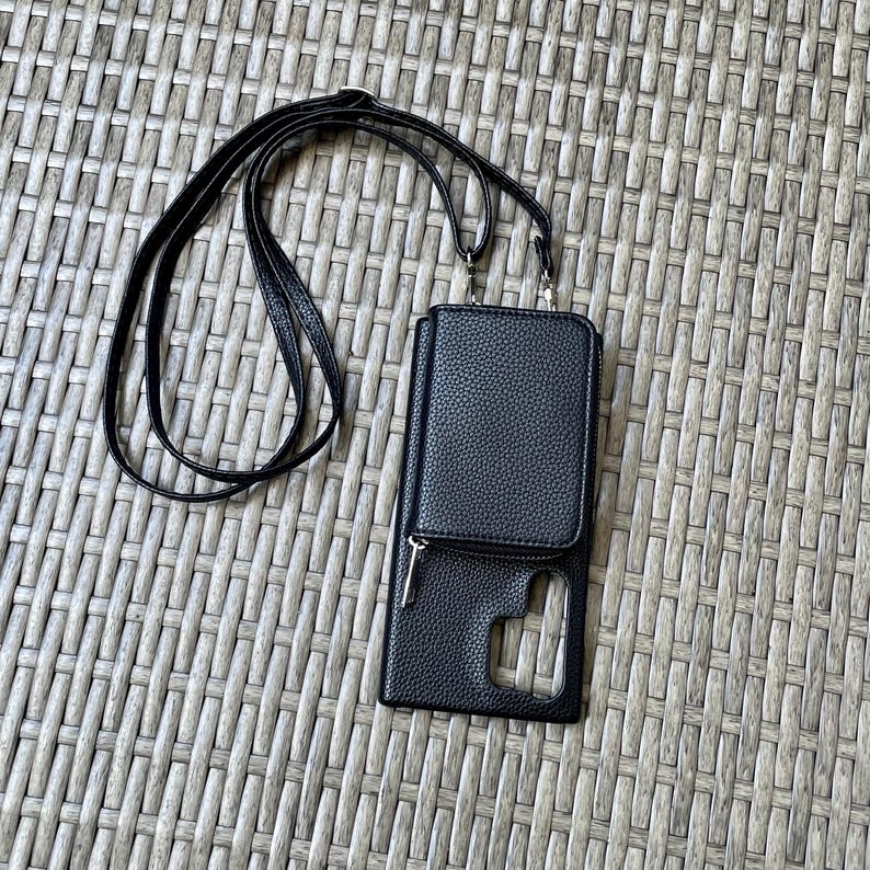 May include: Black faux leather phone case with a zippered pocket and a long black strap. The case has a clear window for the phone screen.
