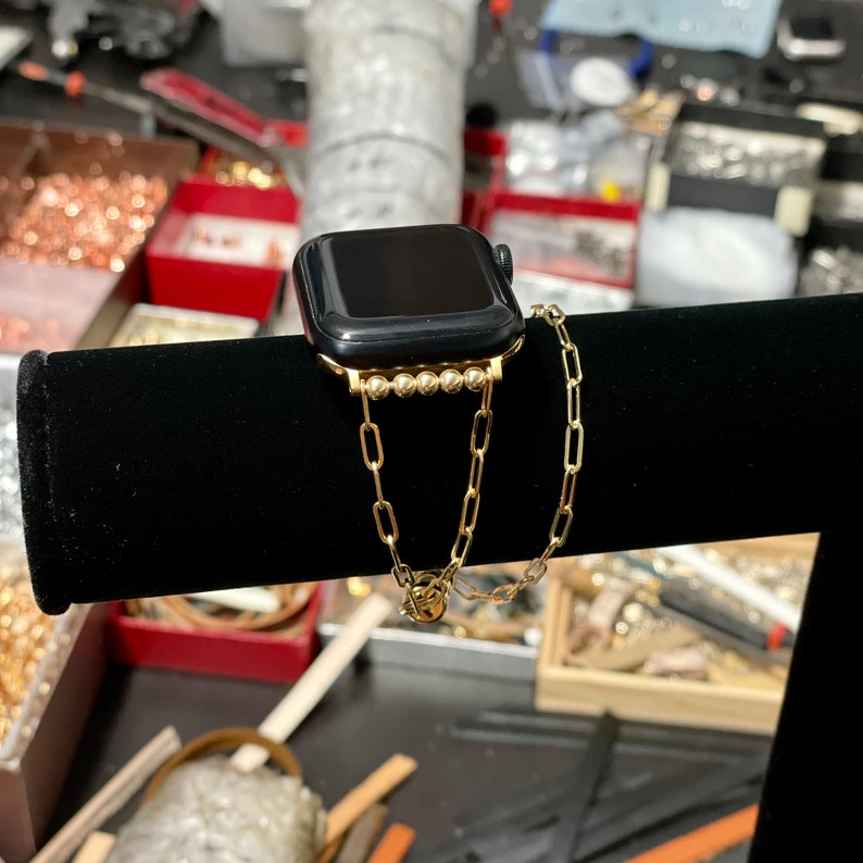 Puede incluir: Una pulsera de reloj de cadena dorada con un Apple Watch negro. La pulsera tiene una fila de peque&ntilde;as cuentas doradas.