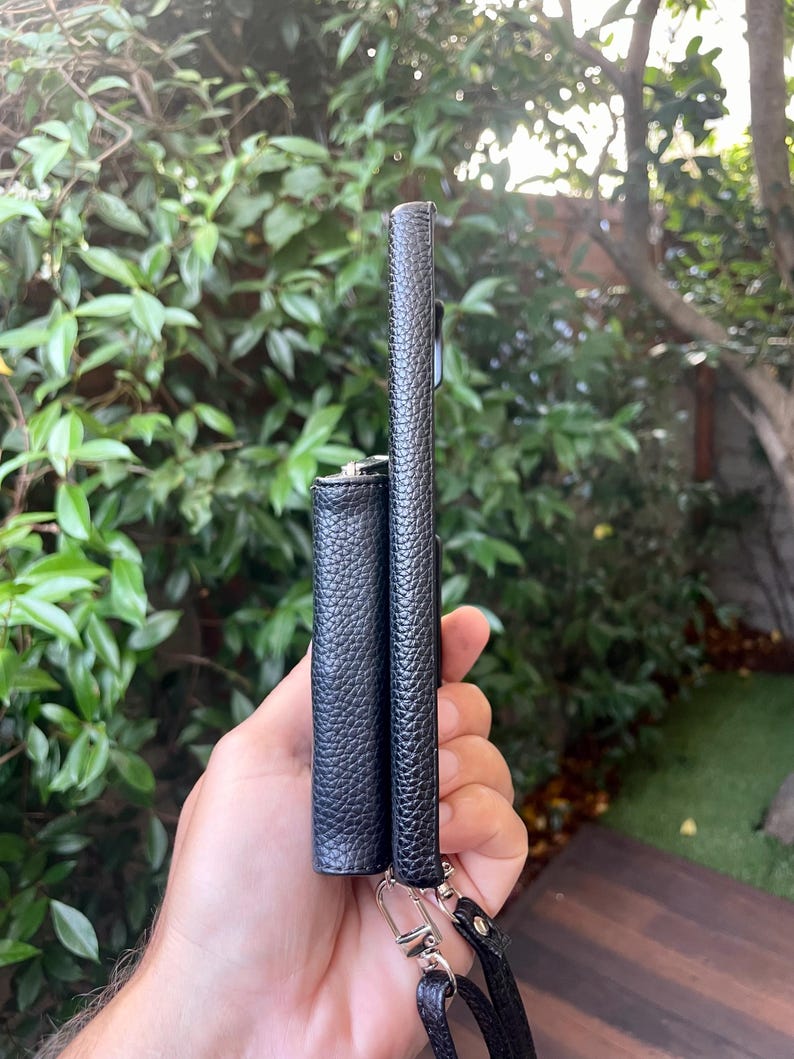 May include: Two black leather phone cases with a black strap. The cases are side by side and have a textured finish.