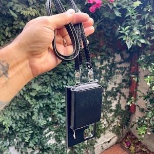 May include: A black leather phone case with a zippered pocket and a black strap. The case is hanging from a hand.