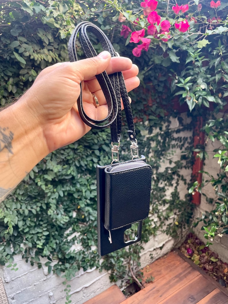 May include: A black leather phone case with a zippered pocket and a black strap. The case is hanging from a hand.