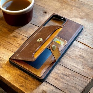May include: Brown leather phone case with a wallet design. The case features a flap with a gold snap closure and a contrasting yellow interior. A small card is visible in a slot. A cup of coffee sits in the background.