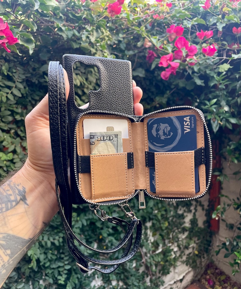 May include: Black leather phone case with a zippered pocket and a strap. The case has a built-in card holder and a cash pocket. The case is open and shows a credit card and cash inside.