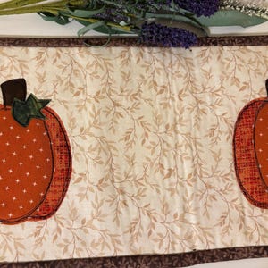 May include: A fall-themed table runner featuring two orange pumpkin appliqués with brown stems and green leaves. The pumpkins are on a cream-colored background with a leaf pattern. The runner has a brown border and is decorated with faux lavender.