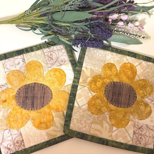 May include: Two square quilted coasters with a sunflower design. The sunflowers have yellow petals and brown centers. The coasters are bordered with green fabric and sit on a white surface. Artificial lavender and greenery are in the background.
