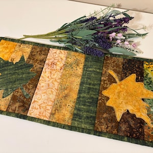 May include: A rectangular table runner with a patchwork design featuring autumn colors. The runner has green and gold leaf appliqués. The background has vertical stripes of brown, gold, and green fabrics. A bouquet of purple flowers rests on the table runner.