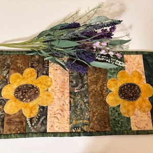 May include: A rectangular quilted table runner with a green border, featuring vertical panels in shades of brown, green, and peach. Two large yellow sunflowers with brown centers are appliqued on the runner. Artificial lavender and greenery are placed on top.