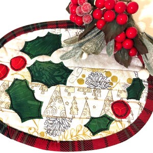 May include: An oval quilted fabric mat featuring a white background with gold-toned line drawings of Christmas trees and pinecones. The mat is bordered by a red and black plaid fabric and decorated with appliquéd dark green holly leaves and red circles.