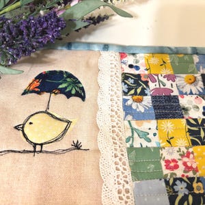 May include: A handmade quilted coaster with a patchwork design featuring floral and geometric patterns in shades of blue, yellow, green, and white. The coaster has a white lace trim and a small embroidered design of a yellow bird holding an umbrella.