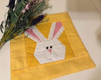 May include: A yellow and white checkered fabric square with a pink and white bunny applique in the center. The bunny has black button eyes.