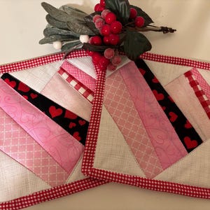 May include: Two pink and red quilted pot holders with a heart design. The pot holders have a white background with a red and white checkered border.