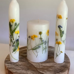 Handmade Unity candles stand on a thick, wooden base. Two long and thin tapers and one lower thick, main candle are white and covered in real flowers. Those flowers are pressed into the candles and they looks like they are painted on the canles.