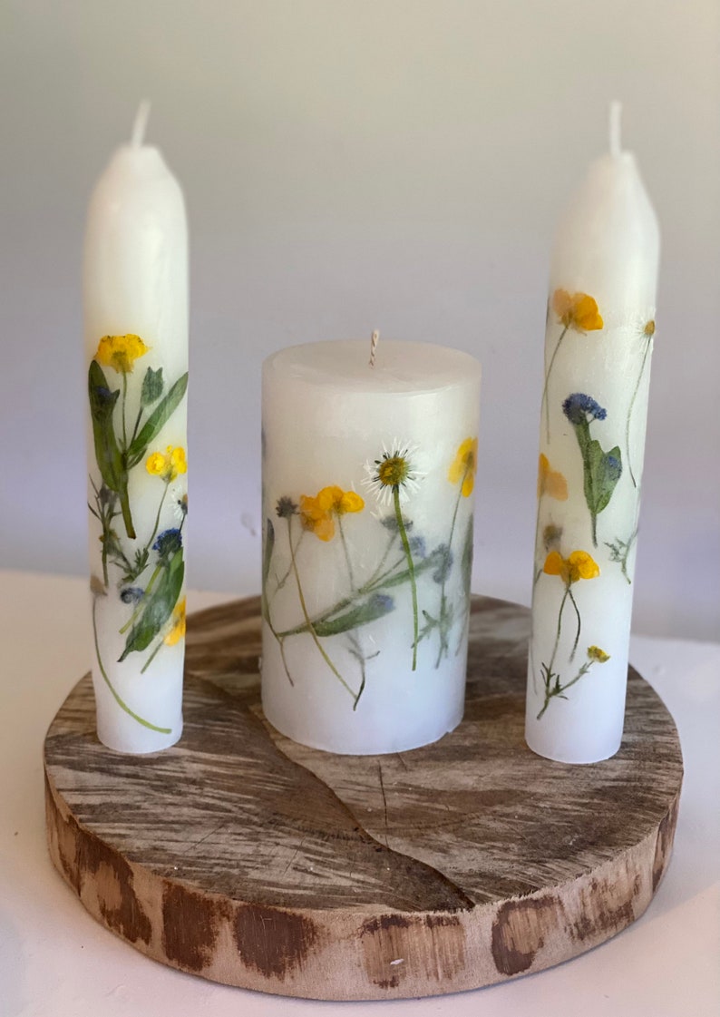 Handmade Unity candles stand on a thick, wooden base. Two long and thin tapers and one lower thick, main candle are white and covered in real flowers. Those flowers are pressed into the candles and they looks like they are painted on the canles.