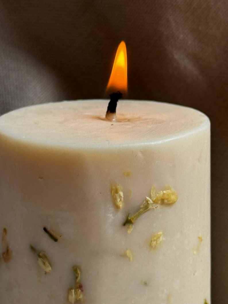 May include: A white candle with a single flame burning at the top. The candle has small, dried flowers embedded in the wax.